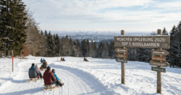 Familien rodeln auf einer verschneiten Rodelbahn in der Umgebung von München mit Blick auf Stadt und Alpenvorland